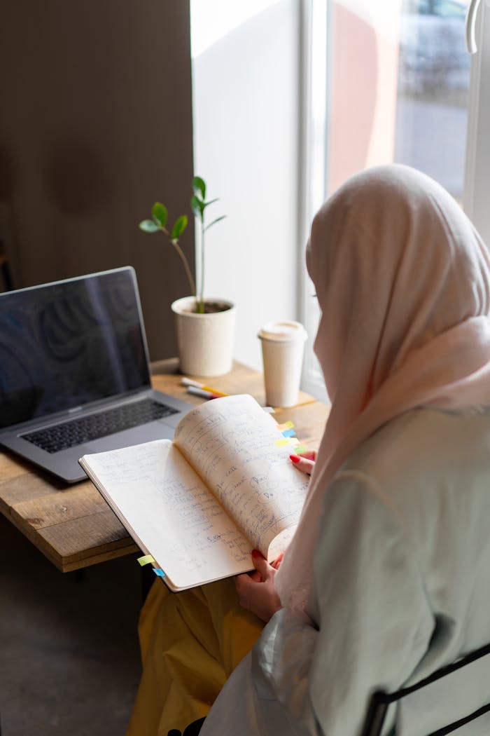 A woman in a hijab reading a notebook by a laptop at a cozy cafe. Perfect for lifestyle and study themes.